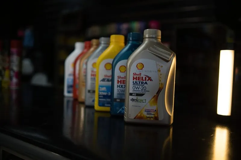 Frasco dourado de Shell Helix Ultra 0W-20 à frente de uma fileira de frascos coloridos, sobre o balcão escuro, com um tubo de luz aceso à direita.
