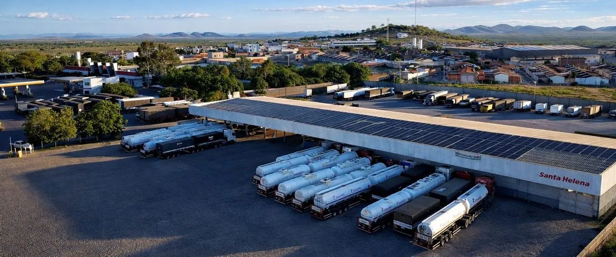 Pátio coberto com placas solares e carretas-tanque estacionadas, visto do alto, com a cidade de Itaberaba e a serra ao fundo.