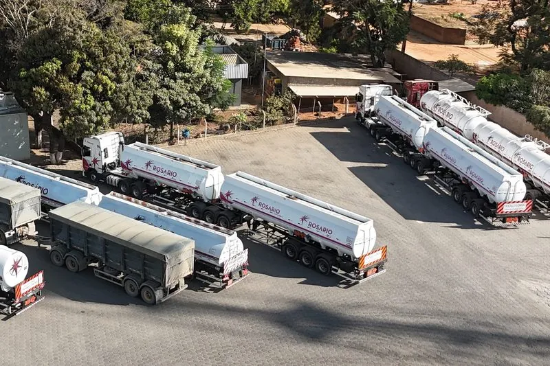 Carretas-tanque Rosário estacionadas em fila no pátio do Posto Rosário, vistas do alto, com uma carreta graneleira de lona no meio e árvores em volta.