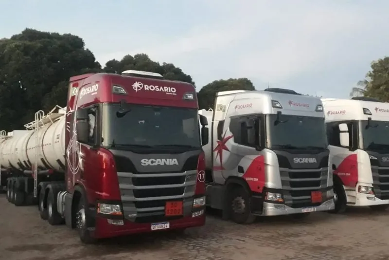 Três cavalos Scania com a pintura Rosário, em vermelho, prata e branco, lado a lado no pátio; o vermelho puxa um tanque.