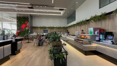 Balcão do Restaurante Premium com a vitrine de sucos e potes de fruta, um frigobar Coca-Cola e o caixa, com plantas em vaso ao lado.