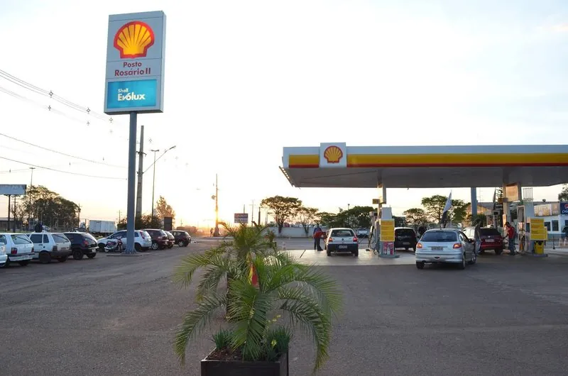 Totem Shell do Posto Rosário II no fim da tarde, com a cobertura e carros abastecendo ao fundo e uma palmeira em vaso na frente.