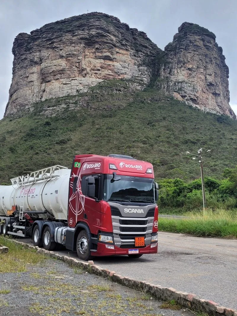 Cavalo Scania vermelho da frota Rosário, puxando um tanque, parado à beira da pista diante de um morro de pedra.